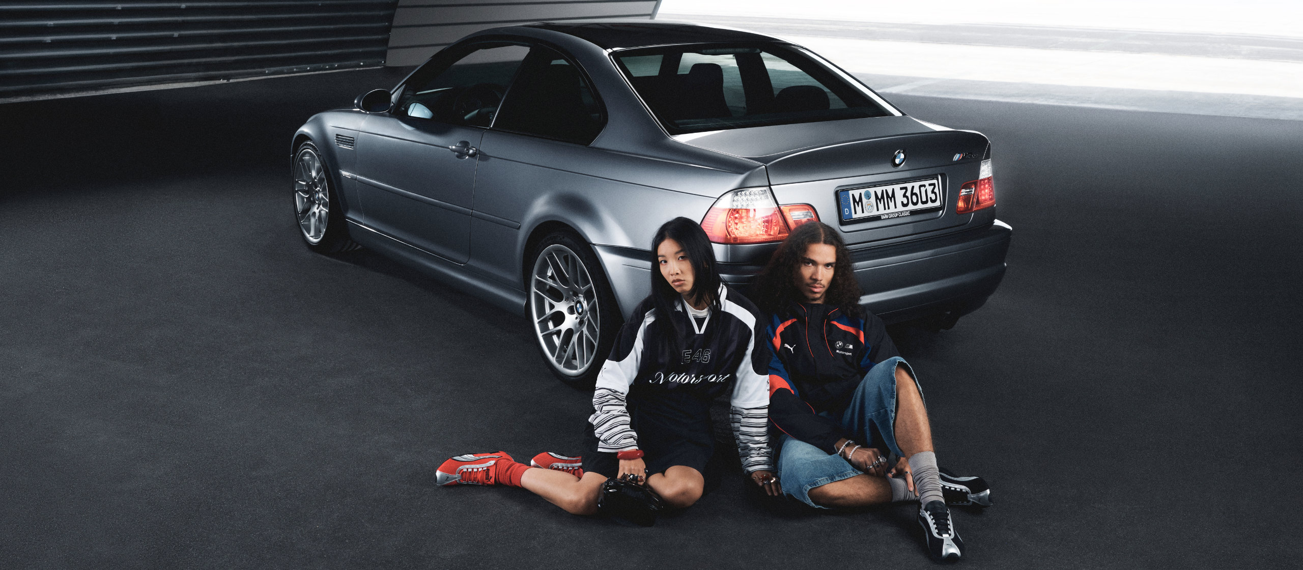A young woman and young man in BMW Motorsport apparel sit behind the rear bumper of a European model BMW 3 Series CSL coupe.